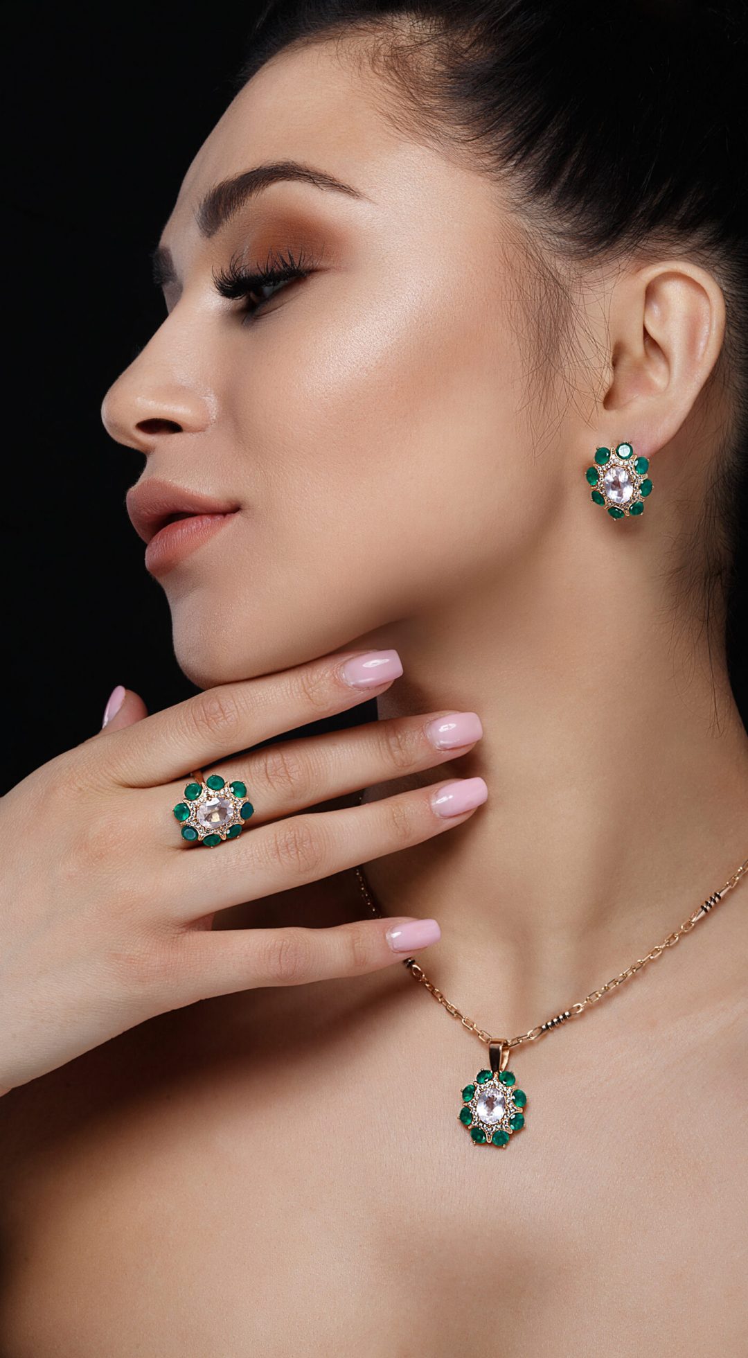 Charming model with dark hair shows rich golden earrings, necklace, and ring with green precious stones